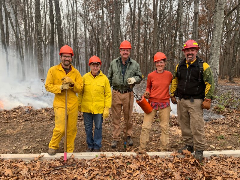 Groups of smiling adults in the woods holding fire tools with a small fire burning behind them. 