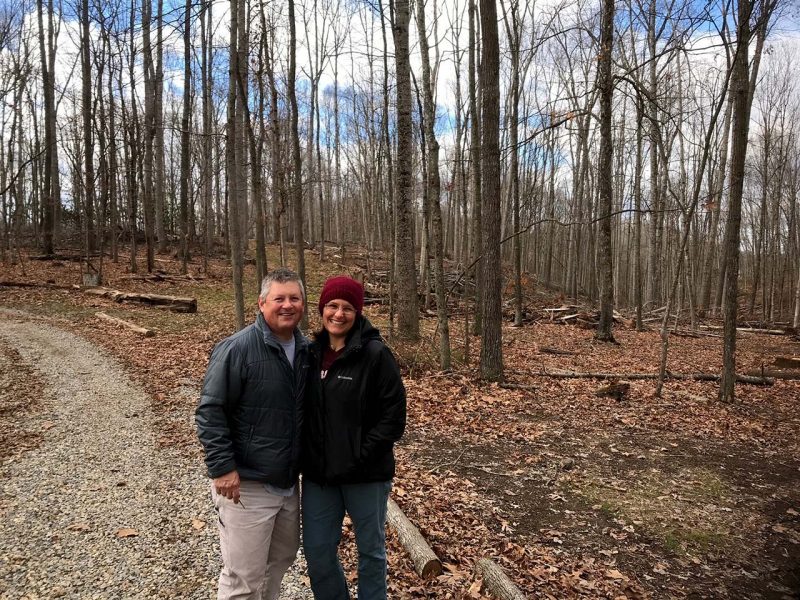 A middle aged man and woman standing in a hardwood forest in the winter.