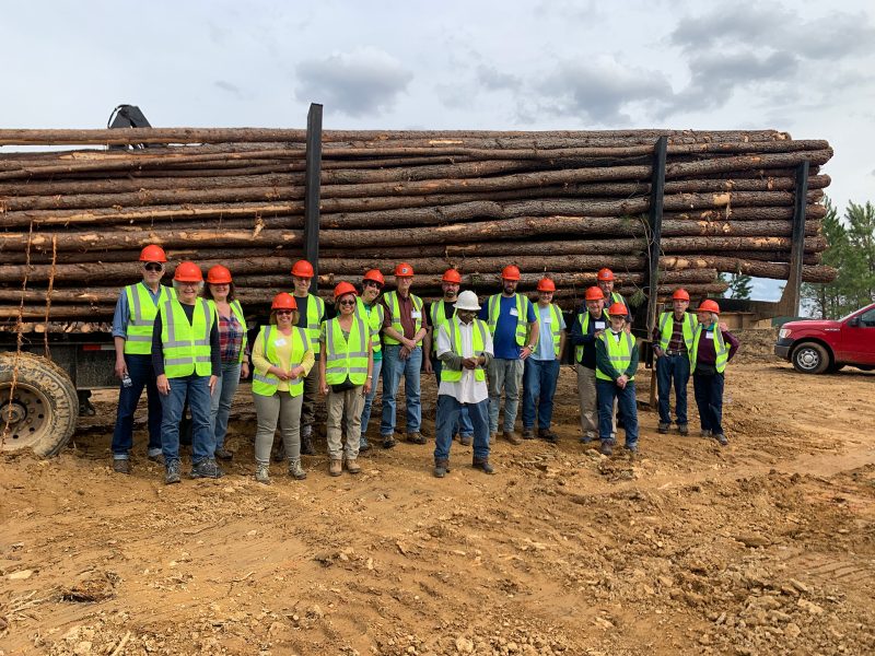 Group of adults standing in front of a loaded logging truck with full-length logs